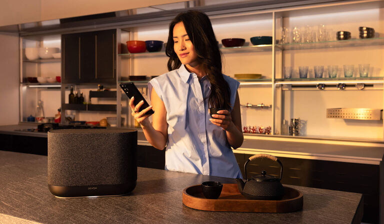 A woman in a kitchen controlling a Denon Home with the HEOS app